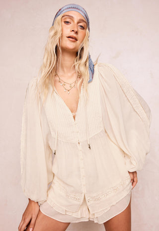 A woman modeling a cream-colored, long-sleeved bohemian-style romper with pleated details and lace trim, paired with a light blue patterned headband, against a neutral beige background.