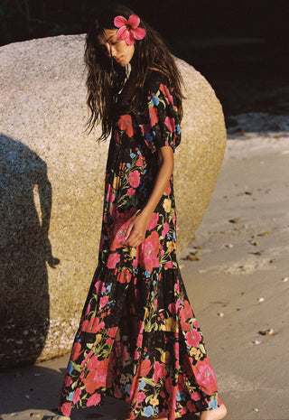 A woman stands on a sandy beach beside a large rock, wearing a long black maxi dress with a vibrant pink, red, and blue floral print. She has a pink flower in her hair.