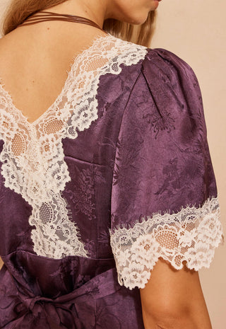 Close-up of the back of a purple satin dress with white lace trim on the shoulders and sleeves, worn by a model.