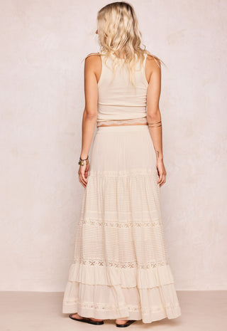 A woman shown from the back wearing a cream-colored sleeveless top and matching tiered maxi skirt with lace trim, standing against a textured beige wall.