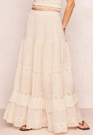A long cream-colored tiered maxi skirt with delicate lace trim and ruffles, shown on a model from the waist down against a light textured wall.