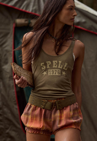 A woman wearing a vintage-style olive green tank top with 'SPELL 1976' printed on the front, paired with plaid shorts, standing in front of a tent.