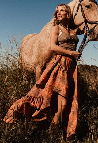 A woman in a rust-orange plaid maxi dress stands outdoors in tall grass beside a light-colored horse, holding its reins under warm golden sunlight.