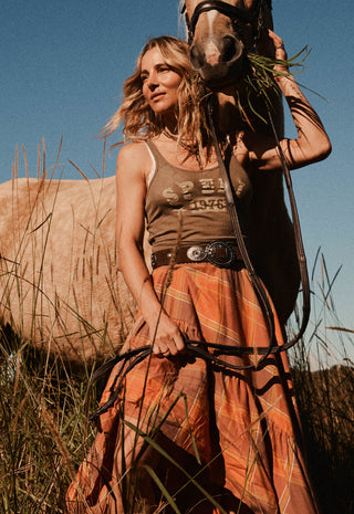 A woman in a rustic western-style outfit stands in a grassy field with a horse beside her. She wears a brown tank top with western text graphics and a long rust-orange plaid skirt, holding horse reins while the horse looks toward the camera under a clear blue sky.