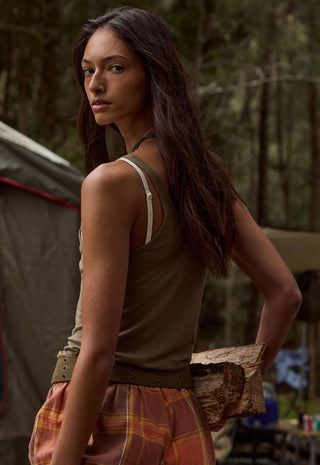 A woman standing outdoors in a forest-like setting, wearing an olive green sleeveless top with white shoulder straps and a brown belt, paired with a rust-orange plaid skirt. She is holding a piece of wood near her hip.