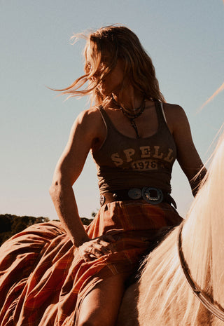 A woman riding a horse outdoors at sunset, wearing a brown tank top with 'SPELL 1976' printed on the front, a rust-orange plaid skirt, and a large silver belt buckle.