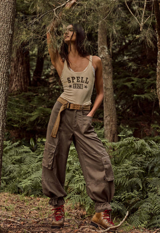 A woman stands in a forest wearing a beige tank top with 'SPELL 1976' printed on the front, loose brown cargo pants, a tan belt, and red hiking boots.