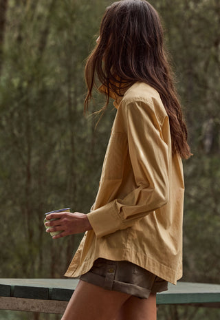 A woman shown from the back wearing a loose, long-sleeve mustard yellow button-up shirt and brown shorts, holding a coffee cup in an outdoor setting with a blurred forest background.