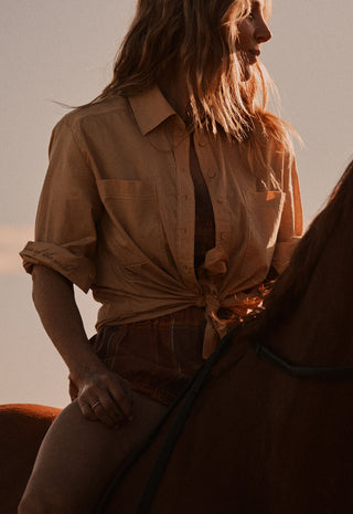 A woman wearing a tan button-up shirt tied at the waist sits on a horse in warm golden-hour light.