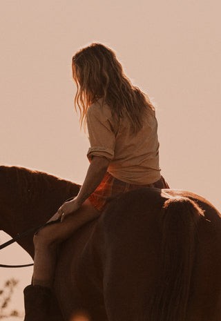 A woman with long light brown hair sits on a horse, shown from behind in warm, backlit outdoor lighting. She wears a beige top and orange plaid shorts against a solid pale peach sky.