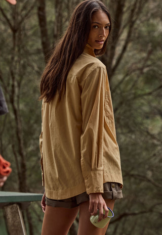 A woman wearing an oversized tan long-sleeve shirt and dark shorts, photographed outdoors against a blurred forest background.