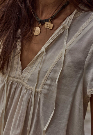 Close-up of a person wearing a loose, short-sleeve bohemian-style top in a light beige or off-white linen-like fabric with lace trim and tie details. The image includes part of the person's face, neck, and hair, and shows a dark cord necklace with wooden pendants.