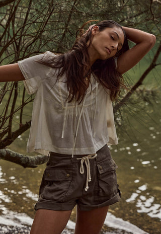 A woman posing outdoors near water and trees, wearing a sheer off-white short-sleeve tie-front top and dark olive cargo shorts.