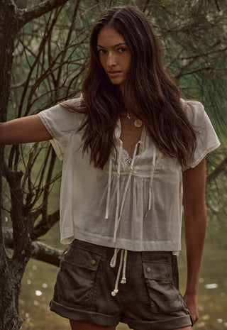 A woman wearing a loose white short-sleeve tie-front blouse and brown shorts, standing outdoors in front of trees.