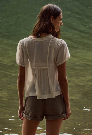 A woman viewed from behind wearing a sheer white short-sleeve blouse and olive green shorts, standing outdoors against a green, water-like background.