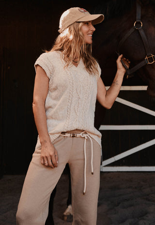 A woman wearing a beige knit sleeveless top, matching beige drawstring pants, and a beige baseball cap stands in a barn holding a horse's bridle. The background shows a dark barn interior with white painted structural lines.