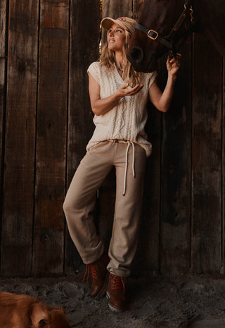 A woman posing in a rustic barn setting with a horse, wearing a cream knit sleeveless top, beige drawstring sweatpants, and brown boots.