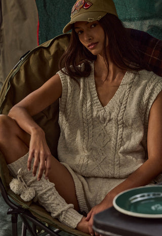 A woman wearing a beige cable-knit sleeveless sweater vest and matching knit socks sits in a camp chair, wearing a khaki baseball cap. A small table in the foreground holds a green plate.