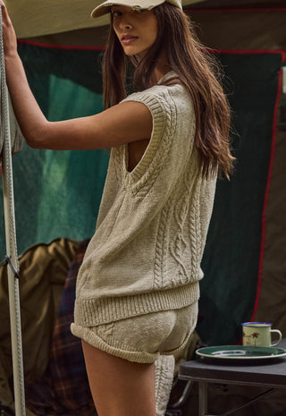 A woman wearing a beige cable-knit sleeveless sweater vest and matching knit shorts, standing in front of a tent with a camping mug on a table in the background.