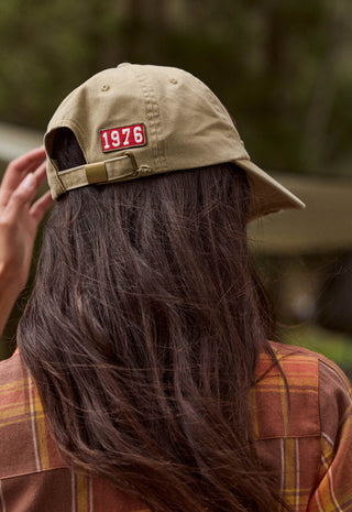 a-person-viewed-from-behind-wearing-a-tan-baseball-cap-with-a-red-rectangular-patch-that-reads-1976-the-person-has-long-dark-brown-hair-and-is-wearing-a-brown-and-orange-plaid-shirt-with-a-blurred-outdoor-background