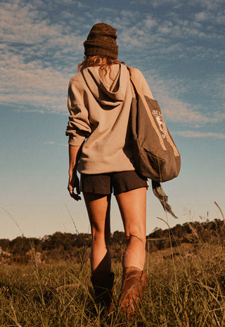 A woman seen from behind walking through a grassy field under a blue sky, wearing a beige hoodie, dark shorts, a brown knit beanie, and carrying a large brown backpack.