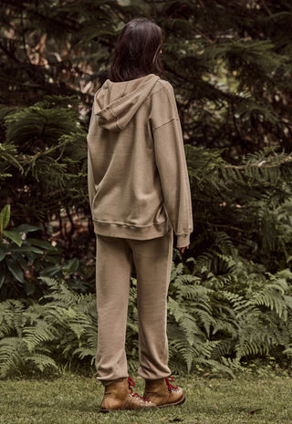 A person standing outdoors in a wooded area, viewed from behind, wearing a matching beige hooded sweatshirt and jogger pants with brown lace-up boots.