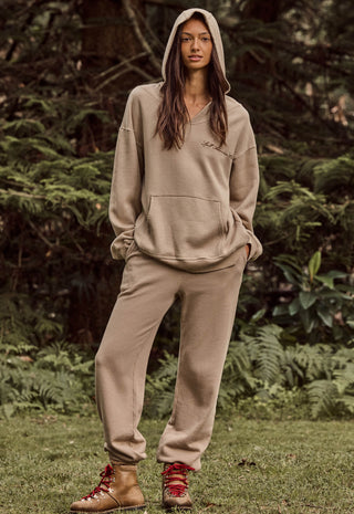 A woman stands outdoors in a wooded setting wearing a matching beige hooded sweatshirt and jogger pants set, with red lace-up ankle boots.