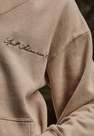 Close-up of a beige crewneck sweatshirt with dark brown embroidered script on the chest reading 'Spell Adventures'.