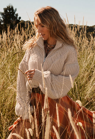 A woman standing in a field of dry golden grass, wearing a chunky cream cable-knit quarter-zip sweater and a rust-orange skirt.
