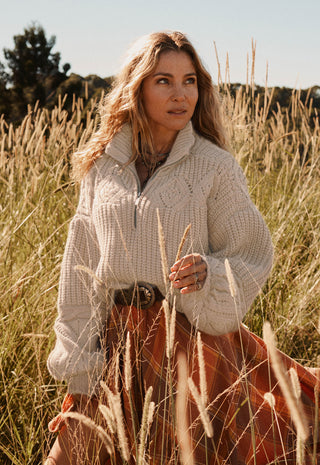 A woman standing in a field of dry grass wearing a cream cable-knit mock-neck sweater and a rust-orange skirt.