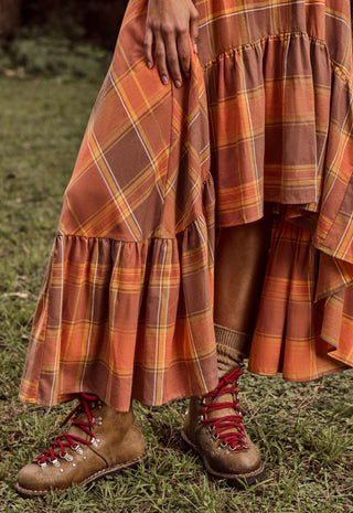 A close-up outdoor shot of a person wearing a long tiered plaid skirt in rust orange, brown, and yellow tones, paired with brown lace-up boots on grass.