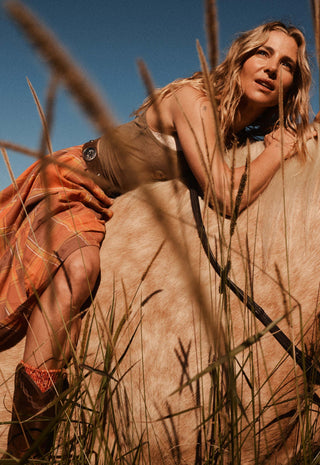 A woman in a desert landscape posing on a light-colored dune with tall grass in the foreground. She is wearing a sleeveless beige top, an orange plaid skirt, and brown boots.