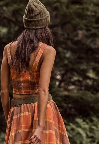 a-woman-viewed-from-behind-wearing-a-sleeveless-rust-orange-plaid-dress-with-a-wide-brown-belt-and-a-matching-knit-beanie-standing-outdoors-against-a-blurred-forest-background