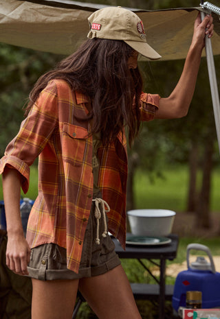 a-woman-wearing-a-rust-orange-and-brown-plaid-flannel-shirt-olive-shorts-and-a-khaki-cap-stands-under-a-tent-at-a-campsite