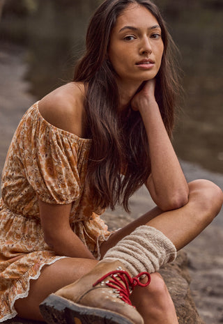 A woman crouching outdoors near water, wearing a loose off-the-shoulder floral dress in warm tan and brown tones, chunky knit socks, and hiking boots with red laces.