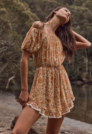 A woman wearing a short off-the-shoulder dress with a rust-orange and beige floral pattern, standing outdoors near water and trees.
