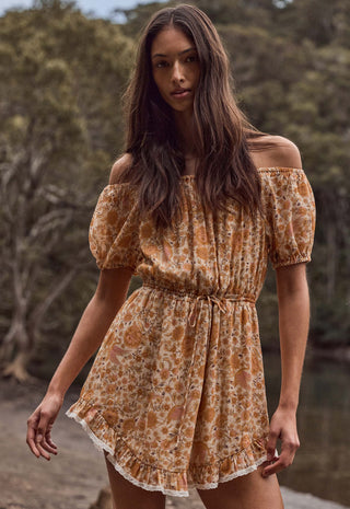 A woman wearing a short, off-the-shoulder dress with puff sleeves and a gathered waist, featuring an orange and beige floral pattern. She is standing outdoors in front of a blurred natural background of trees.