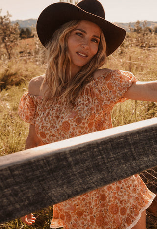 A woman outdoors wearing a black wide-brim hat and a short, off-the-shoulder floral dress in orange and cream tones, standing behind a wooden fence in a sunlit grassy field.