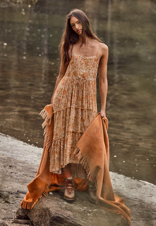 A woman stands outdoors by a body of water wearing a sleeveless, tiered maxi dress with an abstract print in warm tan, beige, and brown tones. She is holding a large rust-orange fringed throw blanket draped over the ground.