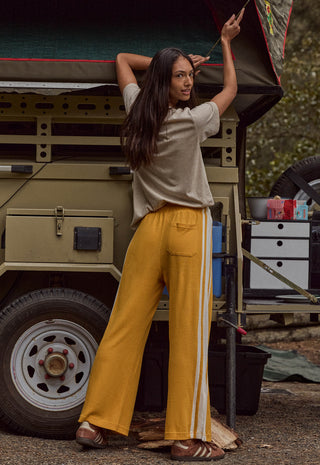 a-woman-stands-outdoors-beside-a-vintage-military-style-vehicle-wearing-loose-mustard-yellow-track-pants-with-white-side-stripes-a-light-beige-short-sleeve-top-and-brown-sandals