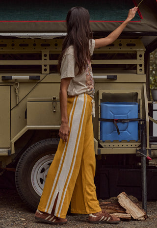 a-woman-stands-in-front-of-a-vintage-military-style-vehicle-wearing-wide-leg-mustard-yellow-pants-with-white-side-stripes-a-beige-graphic-t-shirt-and-brown-sandals