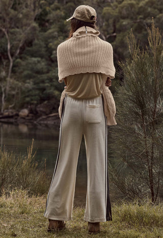 a-person-standing-outdoors-by-a-body-of-water-shown-from-the-back-wearing-a-beige-knit-shrug-beige-top-beige-wide-leg-pants-with-dark-side-stripes-and-a-beige-cap