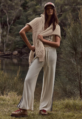a-woman-stands-outdoors-near-a-body-of-water-and-trees-wearing-a-cream-knit-cardigan-draped-over-her-shoulders-cream-wide-leg-pants-with-dark-side-stripes-a-graphic-t-shirt-brown-clogs-and-a-pink-baseball-cap