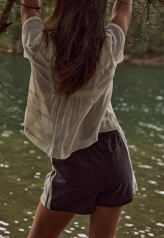 A woman viewed from behind wearing a loose off-white short-sleeve top and dark charcoal shorts with white side stripes, standing outdoors near water and hanging tree branches.