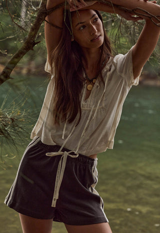 A woman outdoors near water and tree branches, wearing a loose white short-sleeve top and dark charcoal drawstring shorts.