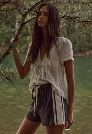 A woman standing outdoors near water and tree branches, wearing a loose white short-sleeve top and dark gray drawstring shorts with white side stripes.