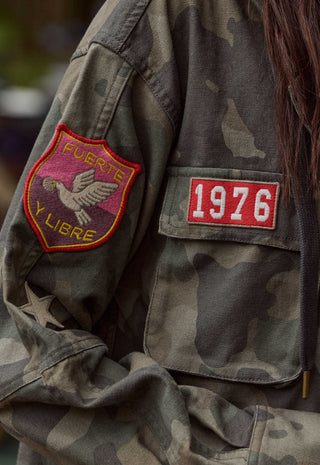 close-up-of-a-camouflage-military-style-jacket-with-a-red-and-yellow-bird-patch-on-the-sleeve-and-a-red-rectangular-patch-with-1976-on-the-chest-pocket