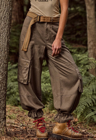 a-person-standing-outdoors-in-a-forest-wearing-loose-brown-cargo-pants-with-gathered-cuffs-a-tan-belt-and-brown-lace-up-boots