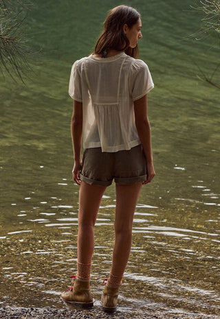 a-woman-stands-at-the-edge-of-shallow-water-viewed-from-behind-wearing-a-loose-white-short-sleeve-blouse-olive-brown-shorts-and-tan-ankle-boots
